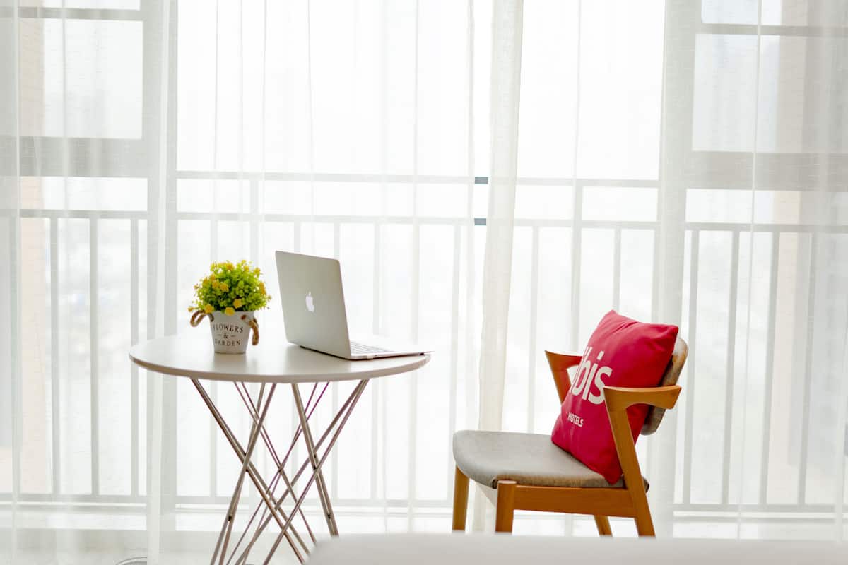 A room with a white table, a laptop, a potted plant, and a chair with a red pillow.