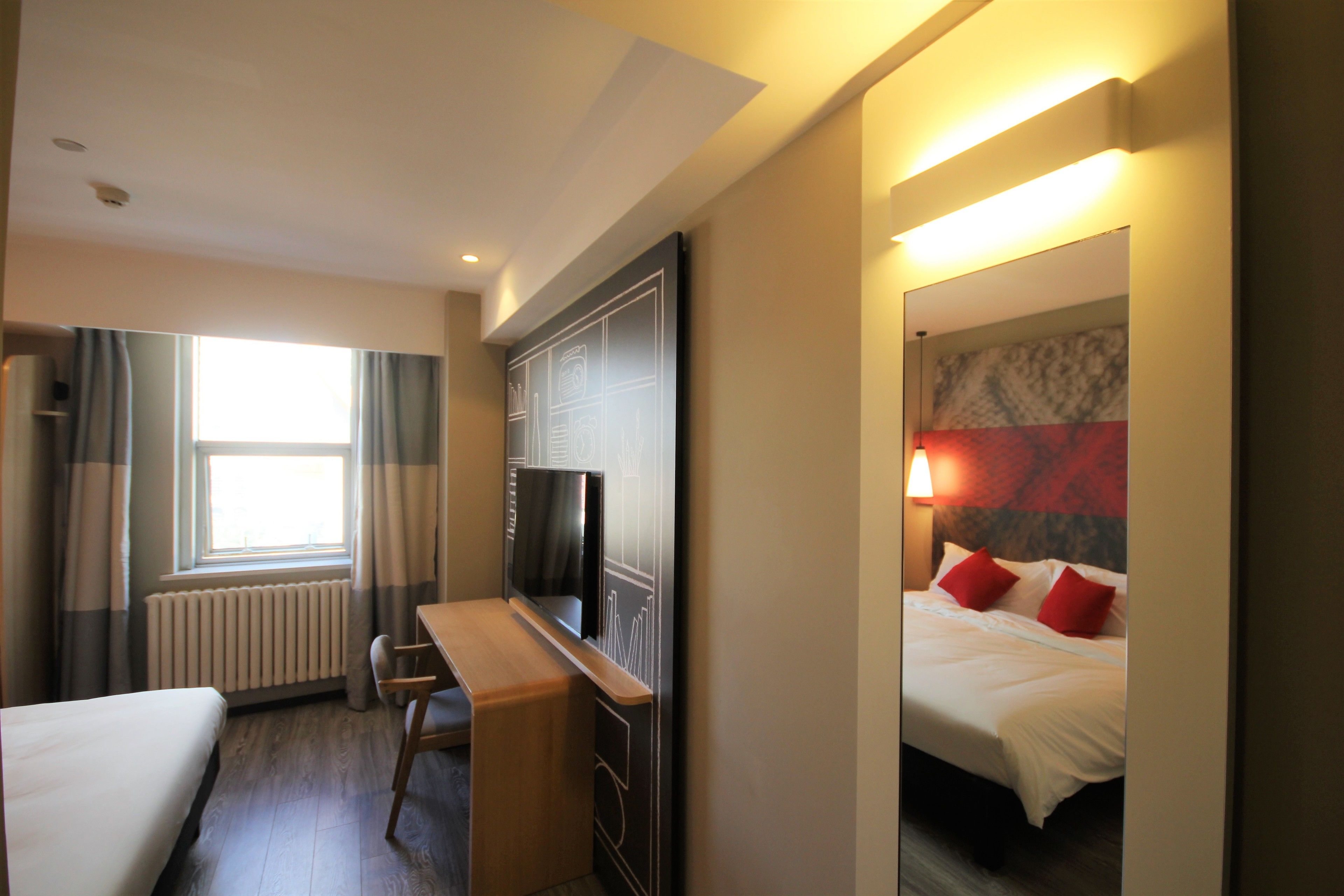 A hotel room with a bed, a desk, a chair, a TV, a window with curtains, and a lamp.