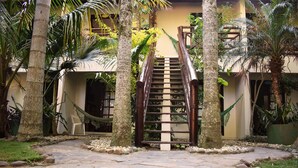 Un escalier en pierre mène à un bâtiment doté d’une balustrade en bois, qui est entouré de palmiers et agrémenté d’un hamac.