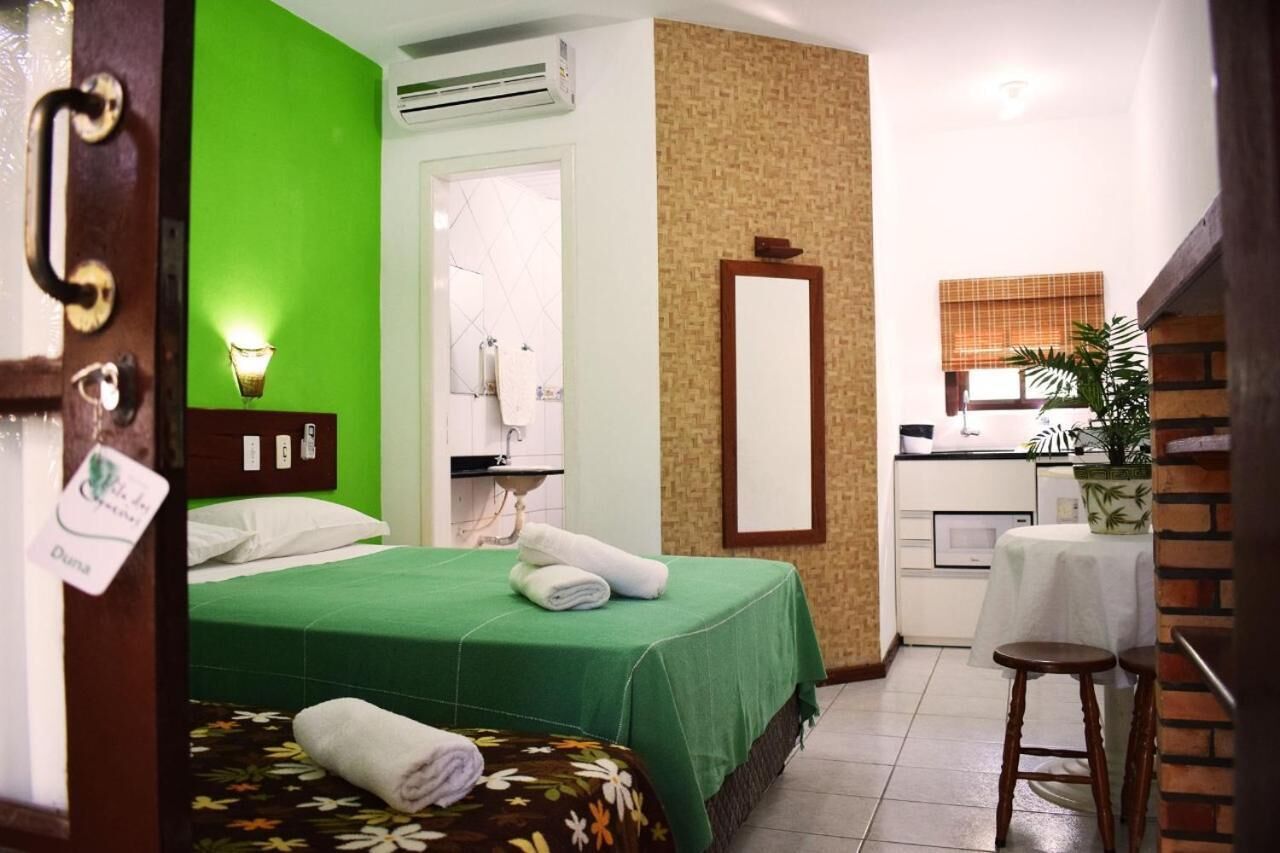 A hotel room with a green bed, a small kitchenette, a bathroom with a mirror, and a potted plant.