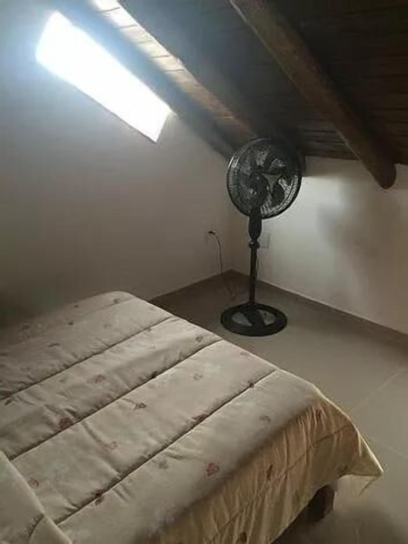 Una habitación pequeña con cama individual, techo inclinado, un ventilador y una ventana que deja entrar luz natural.