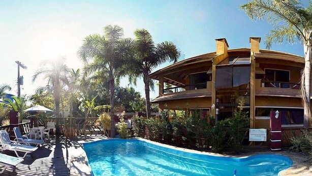 Une piscine entourée de chaises longues et de palmiers, elle-même bordée d’une terrasse en bois et d’un bâtiment à deux étages.