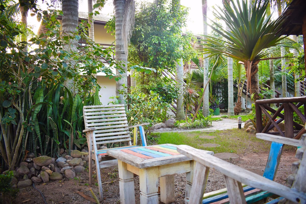 Eine hölzerne Bank und ein Tisch in einem Garten mit tropischen Pflanzen und einem Gebäude im Hintergrund.