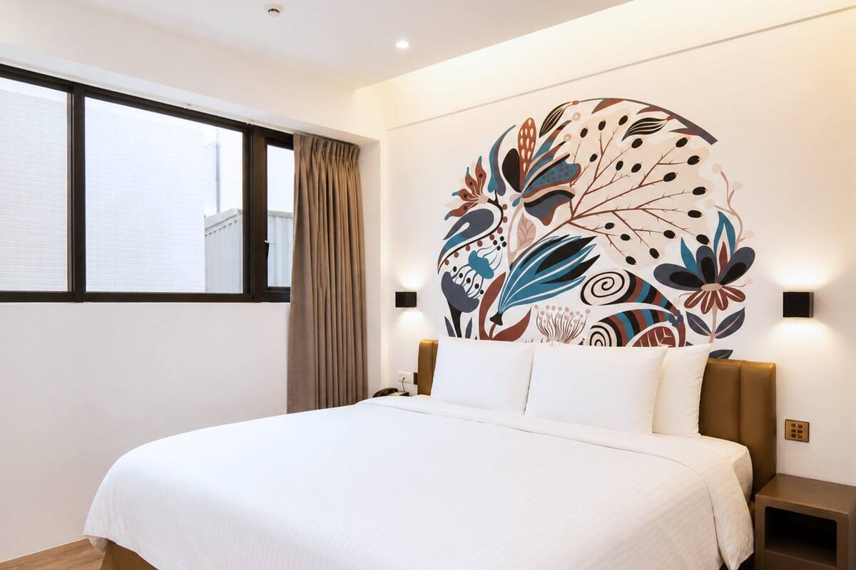 A hotel room with a large bed, a window with curtains, a wall-mounted light, and a decorative wall piece.