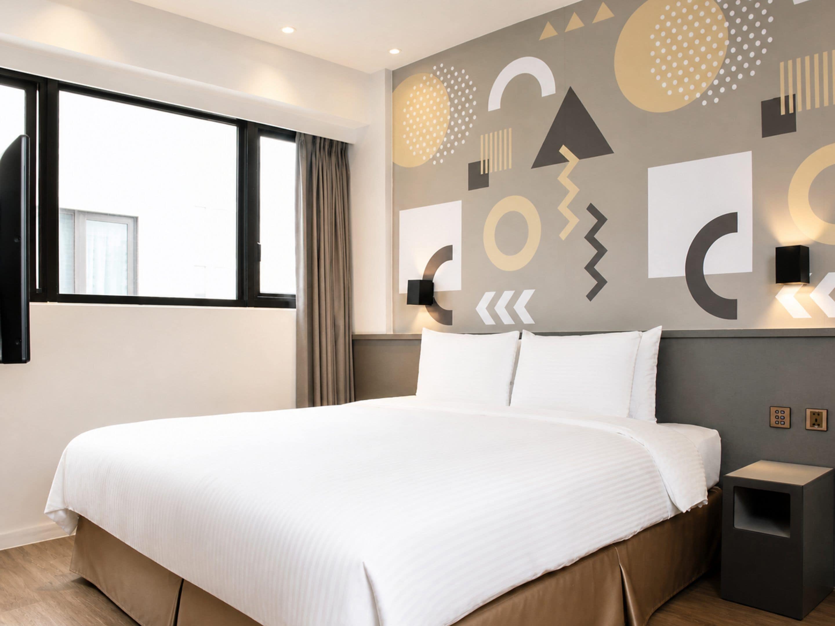 Una habitación de hotel moderna con una cama grande, una ventana con cortinas y un diseño geométrico en la pared.