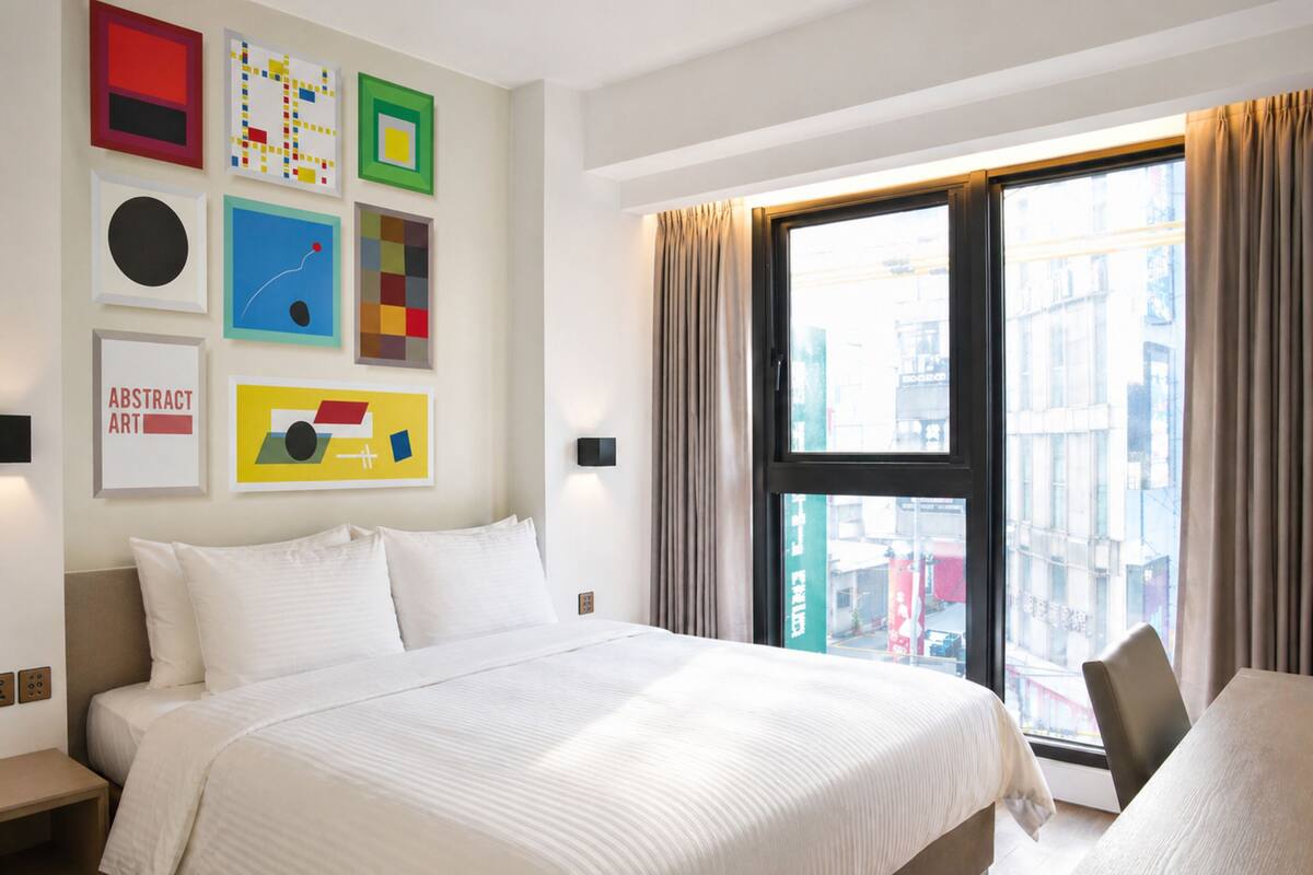 A modern hotel room with a large bed, a desk, and a city view through the window.