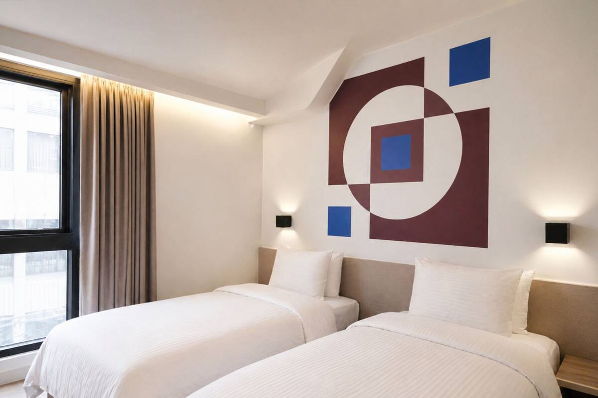 A hotel room with two beds, a large window with curtains, and a geometric wall art piece.
