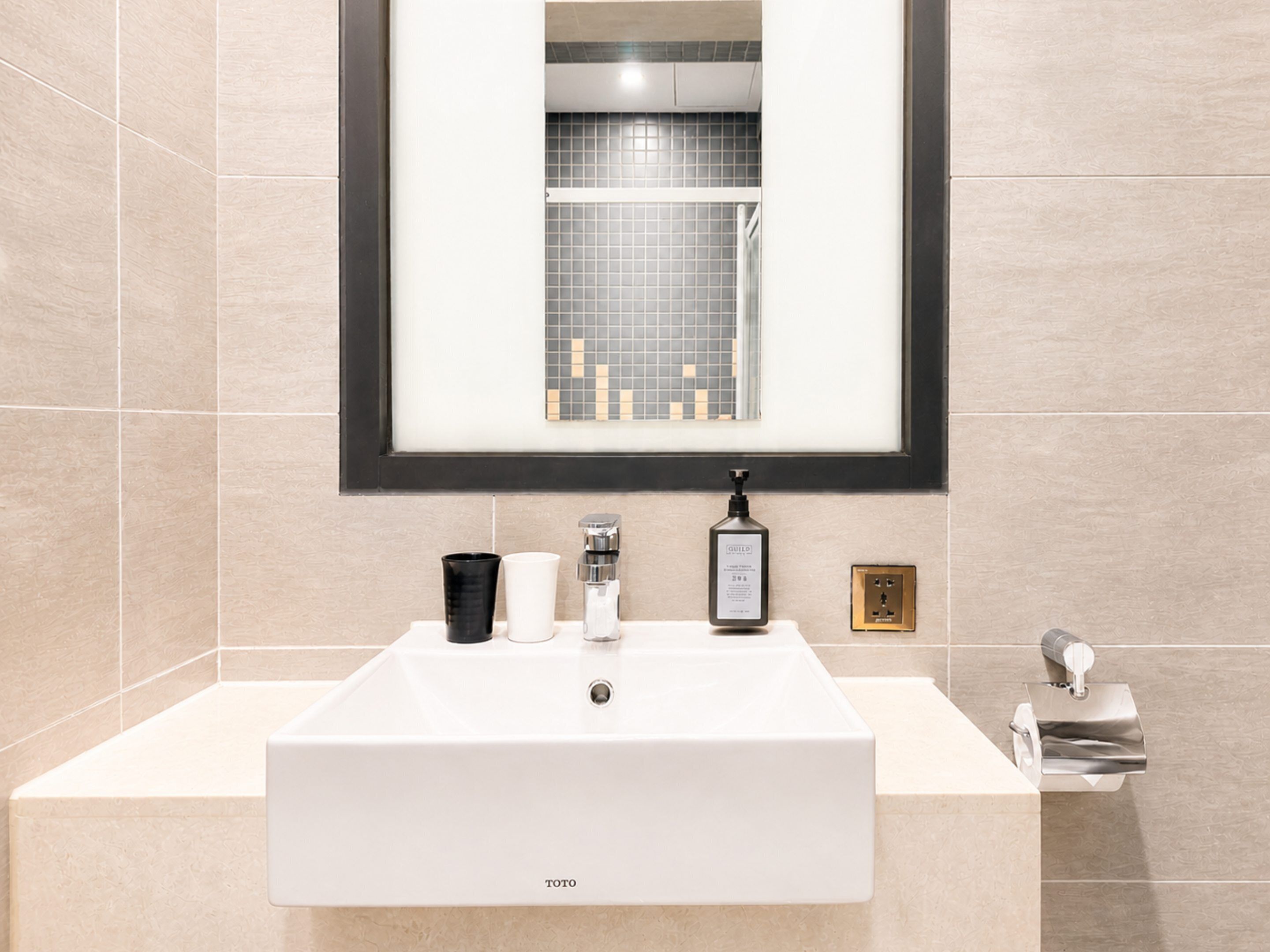 A bathroom with a white sink, a framed mirror, a black soap dispenser, and a small framed picture on the wall.