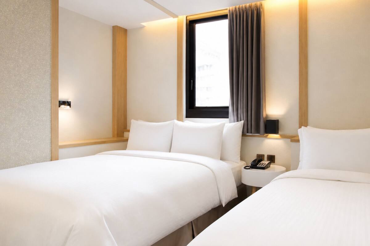 A hotel room with two beds, a window with curtains, a bedside table with a phone, and a wall-mounted light.