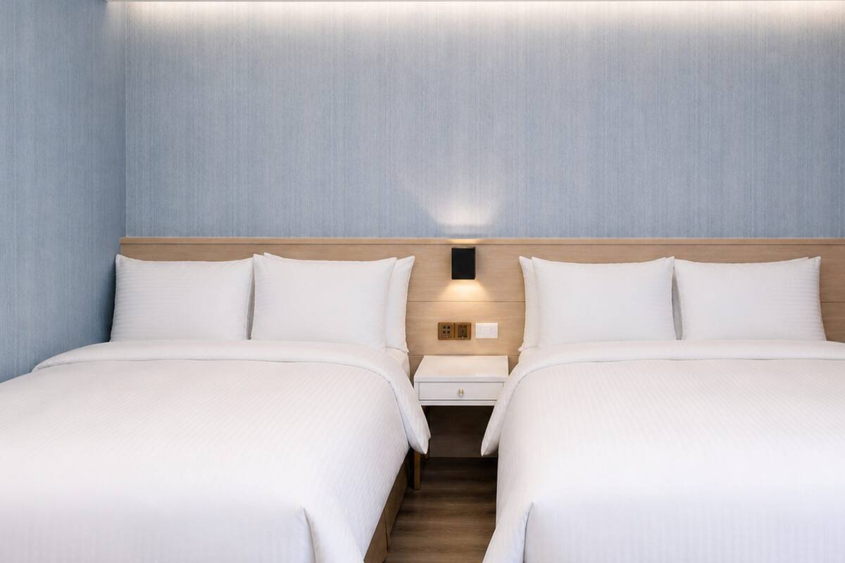A hotel room with two beds, a wooden headboard, a nightstand, and a wall-mounted light.