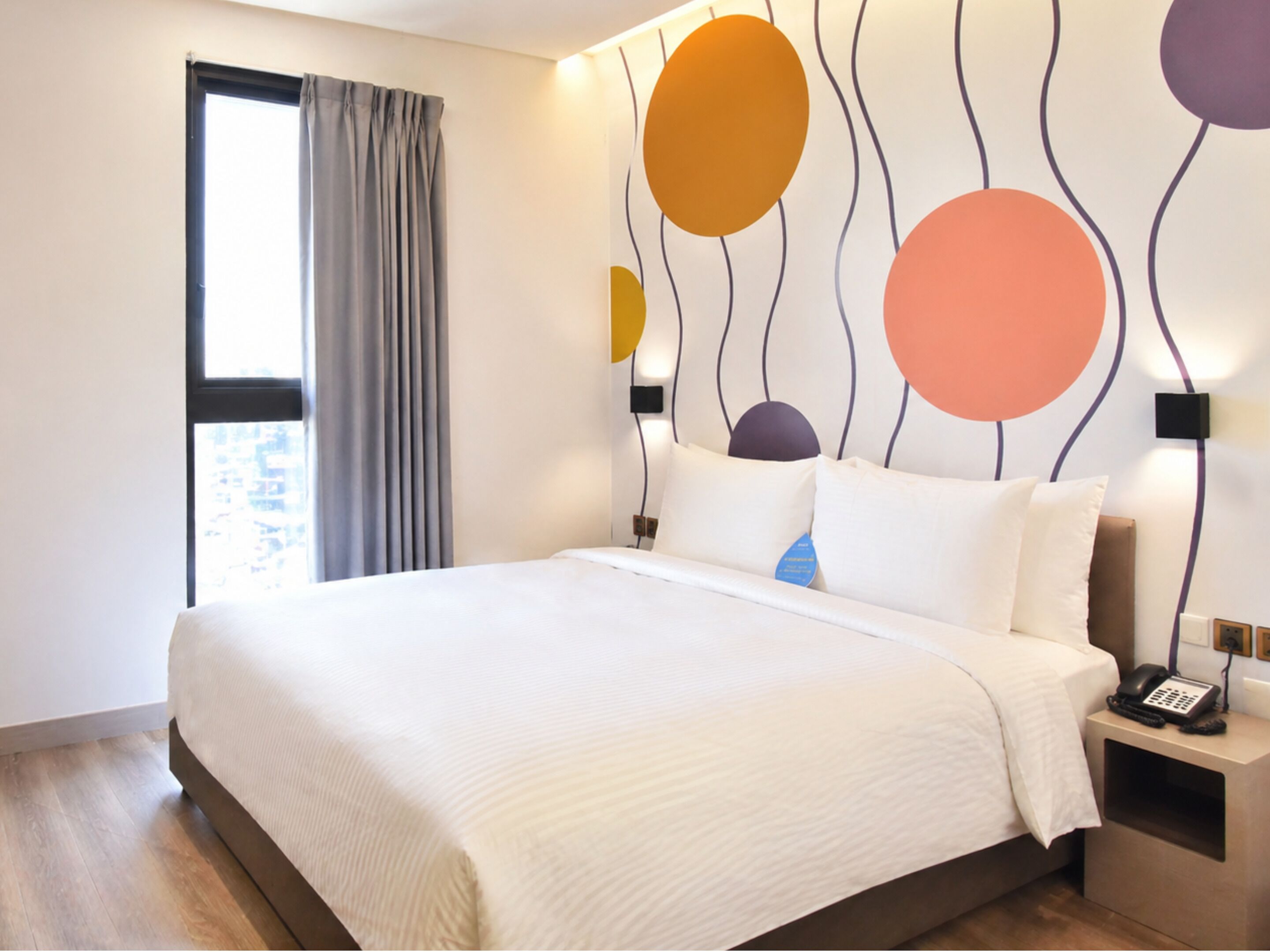 Ein Hotelzimmer mit einem großen Bett, einem Nachttischtelefon, einem Fenster mit Vorhängen und einer Wand mit abstrakten Kreismustern.