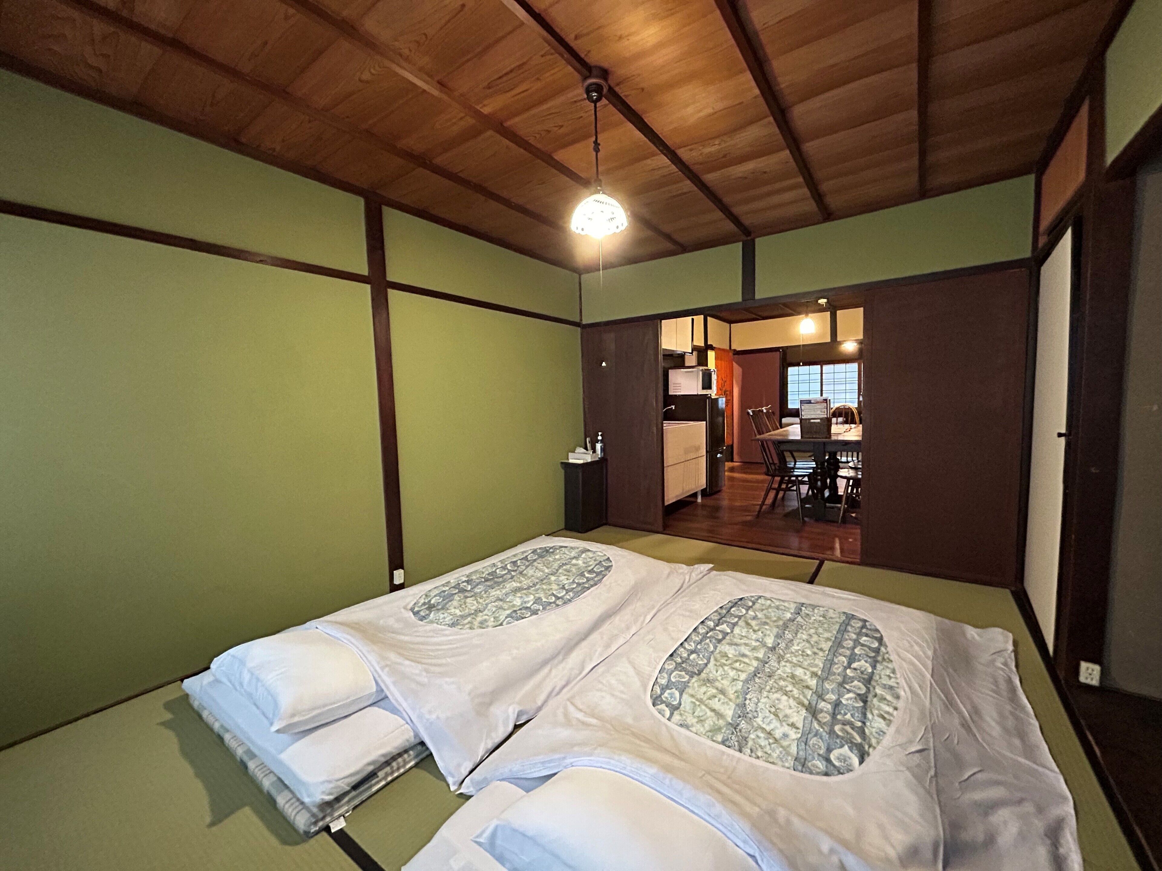A room with two beds, a wooden ceiling, a small kitchen area, and a view of the outside.