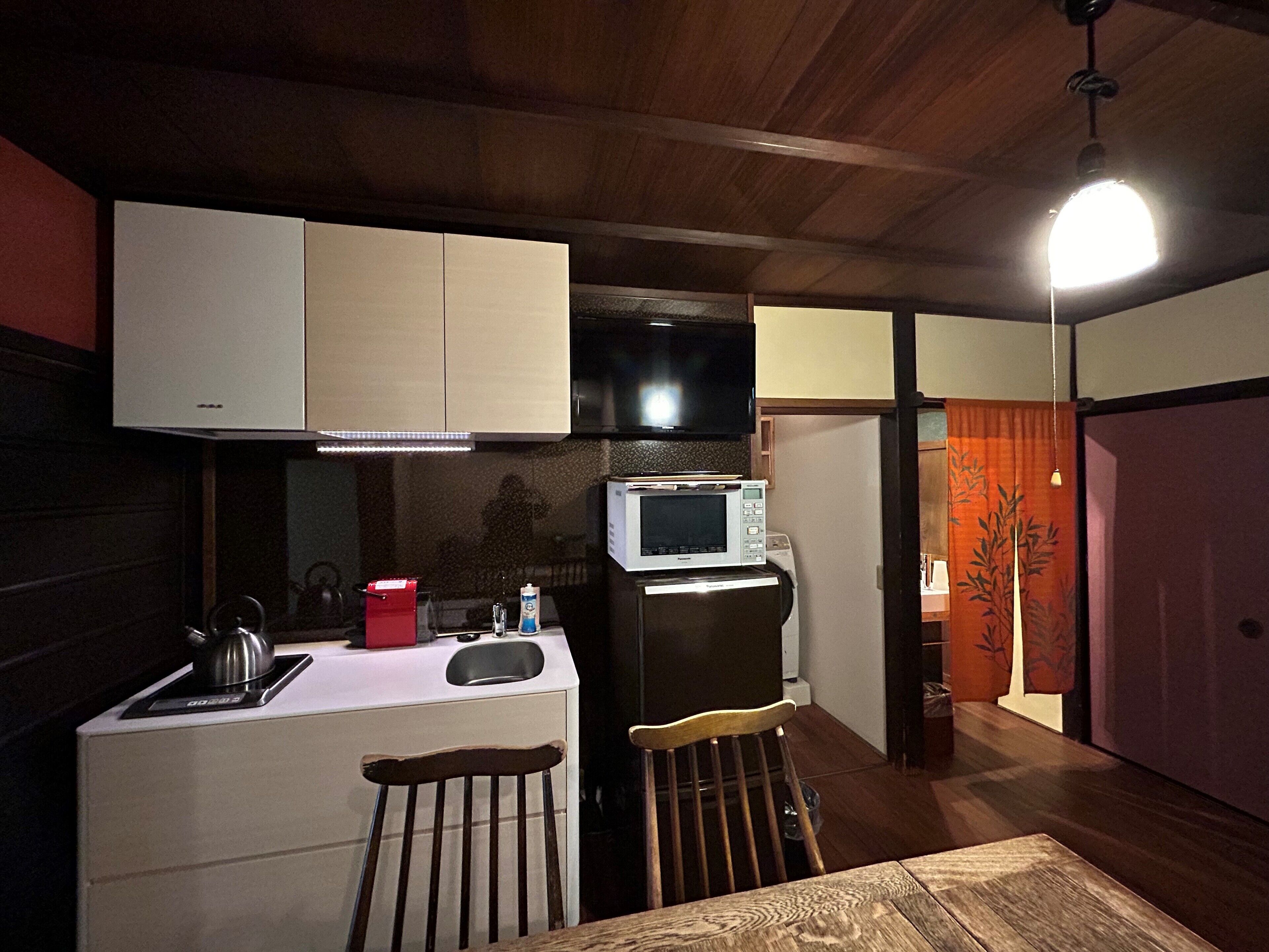 A compact kitchen with a microwave, stove, and sink. There is a wooden chair and a table in the foreground.