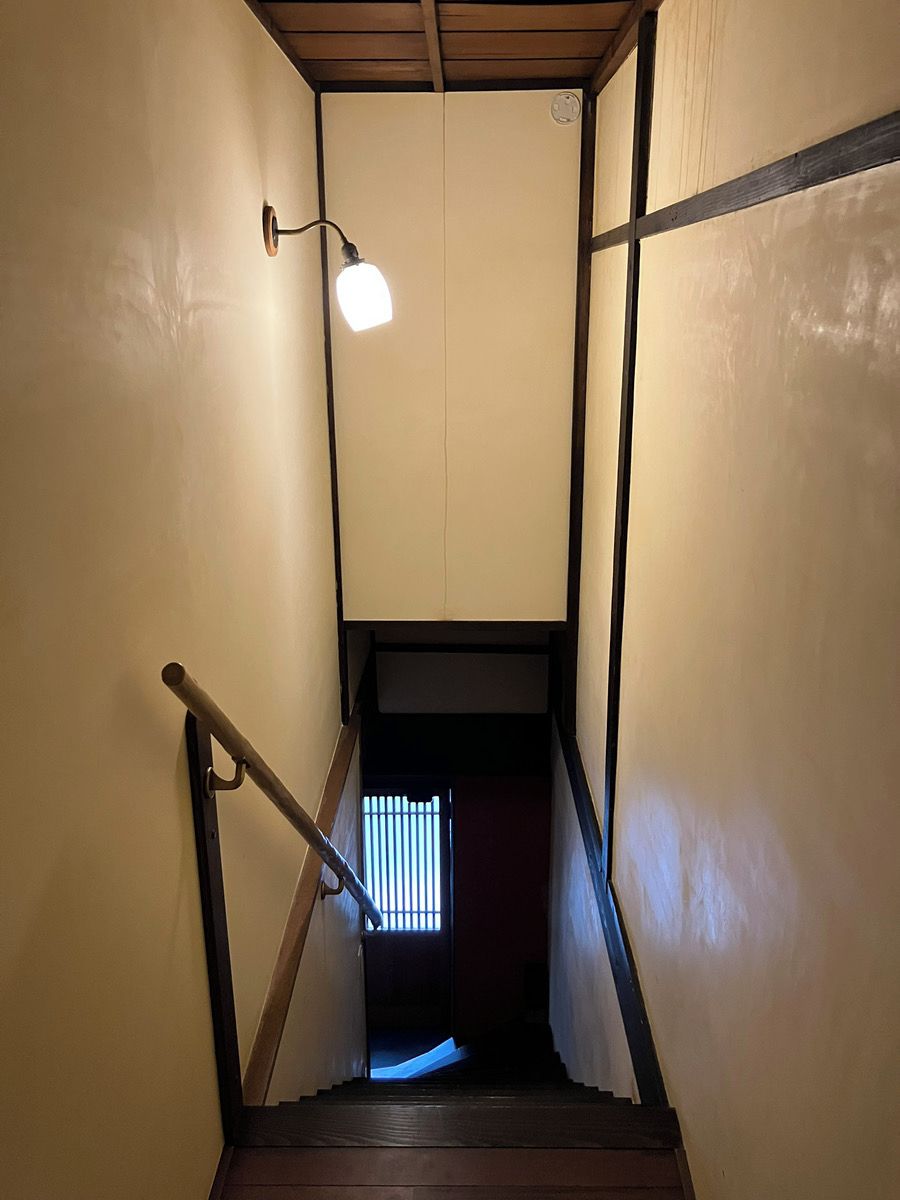 A narrow stairway with a handrail, a light fixture, and a window at the bottom.