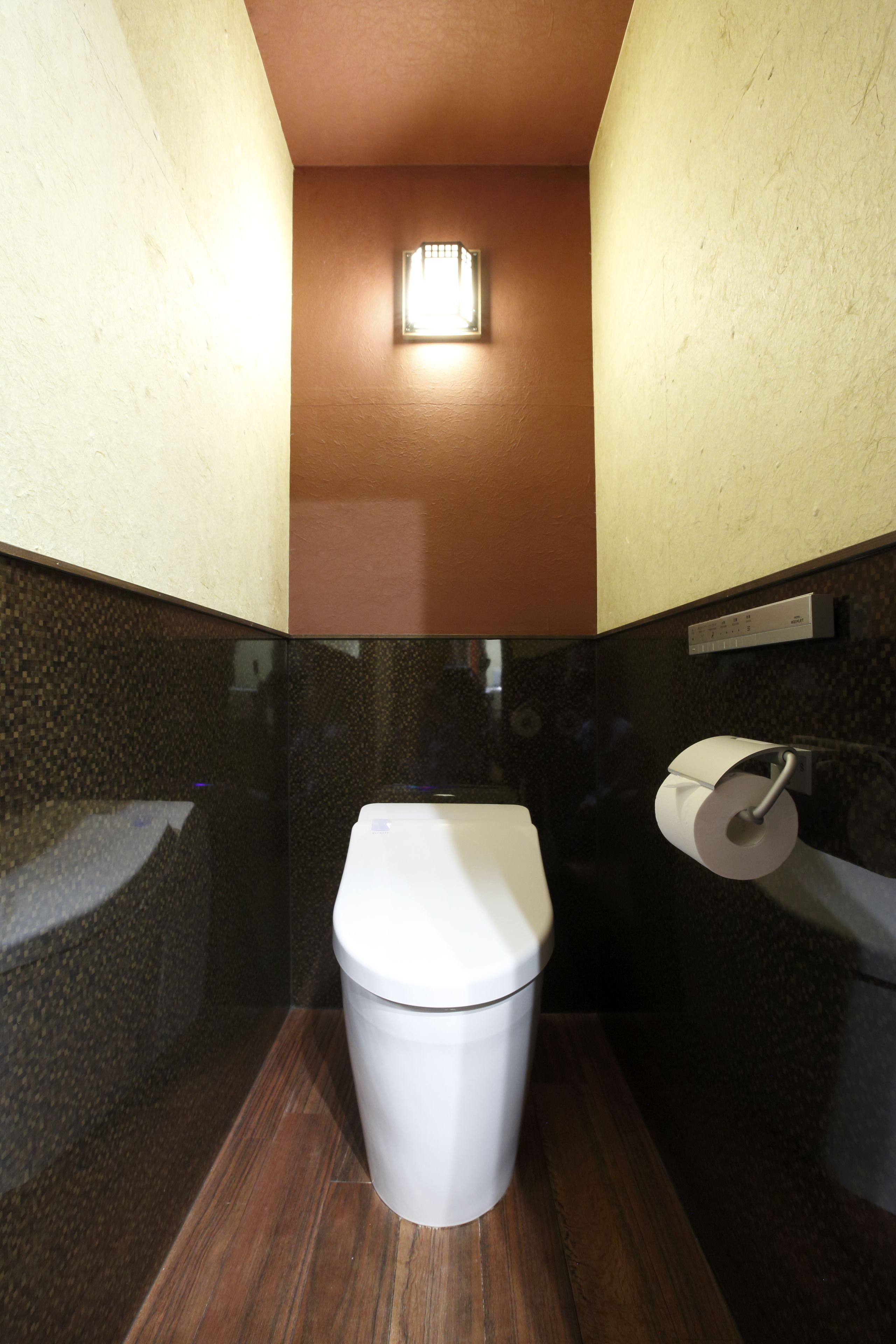 A compact bathroom with a toilet, a paper towel dispenser, and a wall-mounted light fixture.