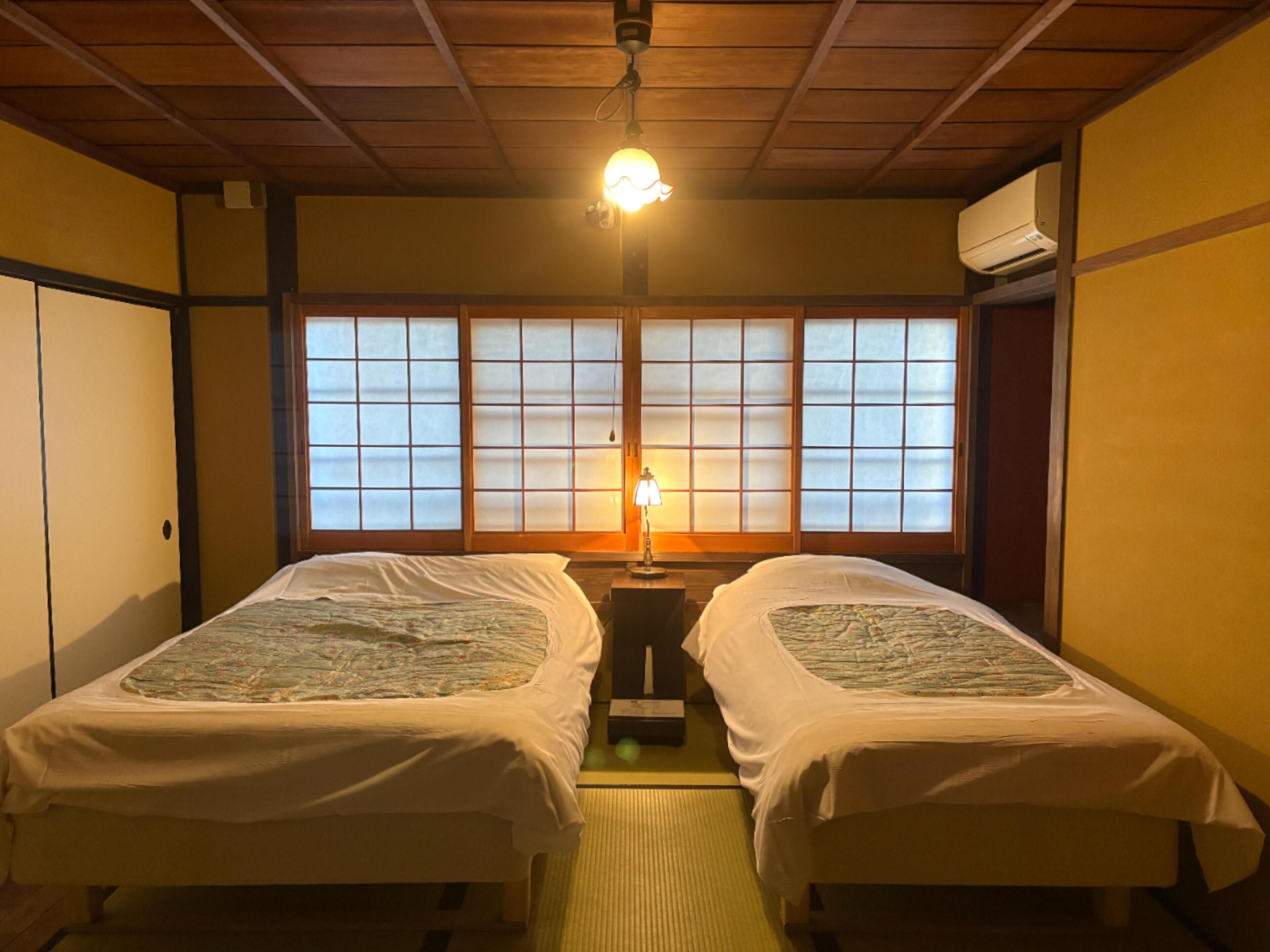 A room with two beds, a window with wooden frames, a lamp on a bedside table, and an air conditioning unit on the wall.