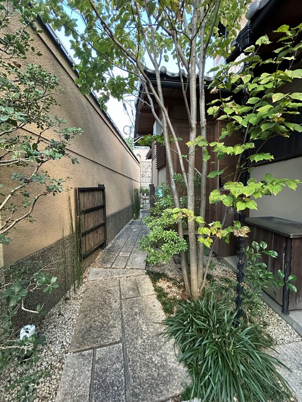 A narrow stone pathway flanked by a building with a wooden gate and a tree with green leaves.
