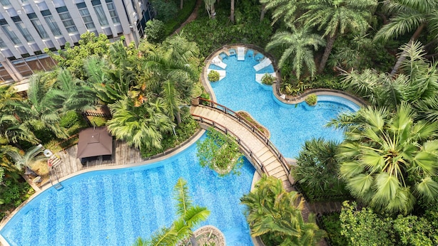 Une grande piscine à plusieurs niveaux, entourée d’une végétation luxuriante et de palmiers, avec un bâtiment moderne en arrière-plan.