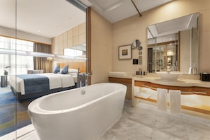 Una habitación de hotel moderna con una bañera grande, una cama doble con ropa de cama azul, un espejo amplio y un baño con lavabo y tocador.