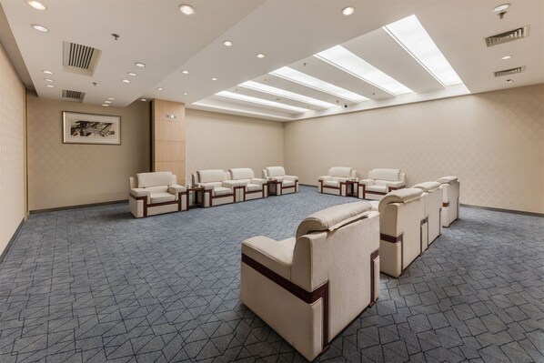 Una habitación amplia con techo alto, iluminación empotrada y tragaluz. Cuenta con una hilera de sofás y sillones beige dispuestos en semicírculo, y un piso con diseño.