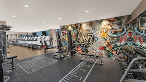 Une salle de sport équipée de tapis de course, d’appareils de musculation et ornée d’une fresque murale aux couleurs vives.