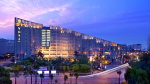 Un hôtel moderne doté d’une façade verticale imposante, de fenêtres illuminées et d’un environnement bien entretenu, agrémenté de palmiers et bordé d’une rue animée.