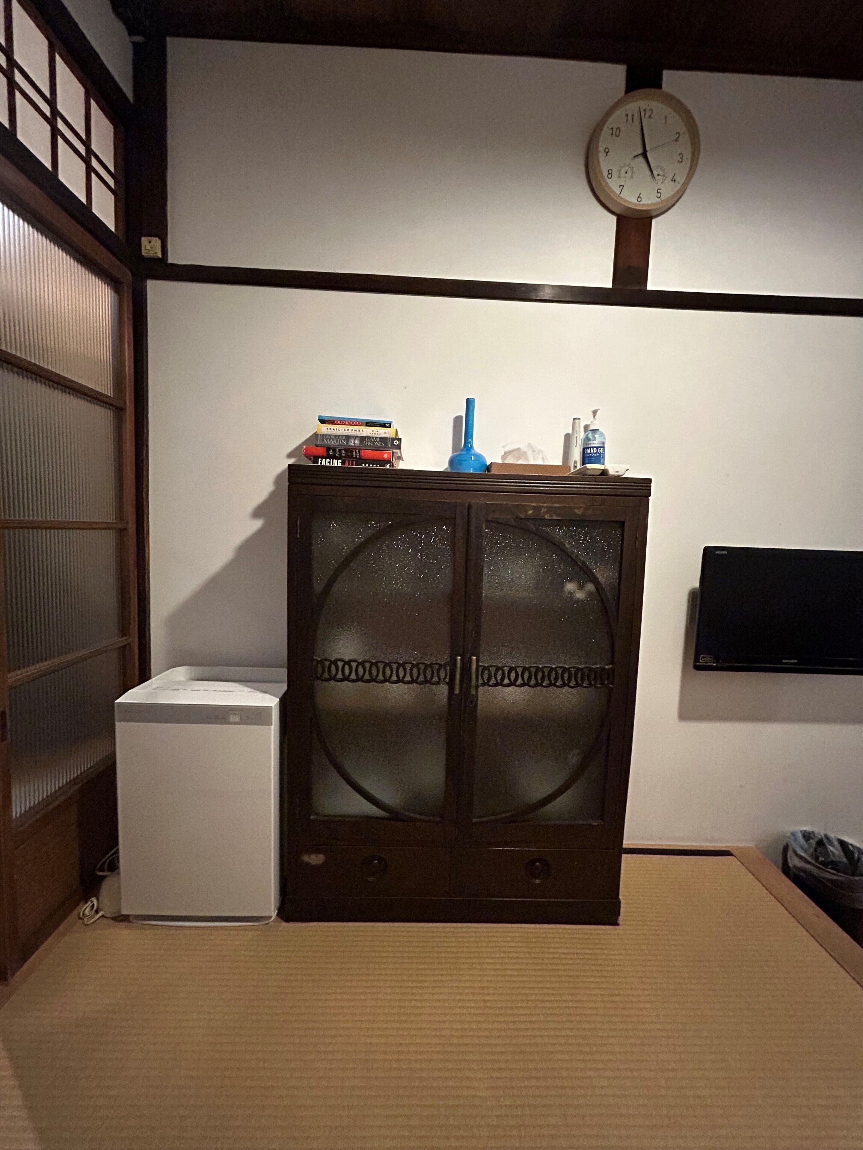 A room with a traditional Japanese-style cabinet, a small white appliance, a clock, and a television on the wall.