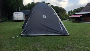 A black tent with a circular logo on the side, set up on a grassy lawn with a trailer and a building in the background. - Guest House Coco Garage - Hostel (Kirishima)