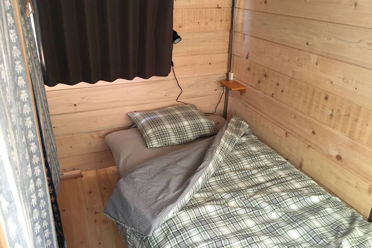 A small, wooden room with a single bed, a pillow, a blanket, and a small shelf.