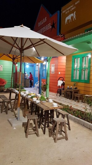 An outdoor dining area with wooden tables and benches, a white umbrella, and a building with colorful walls and signage.