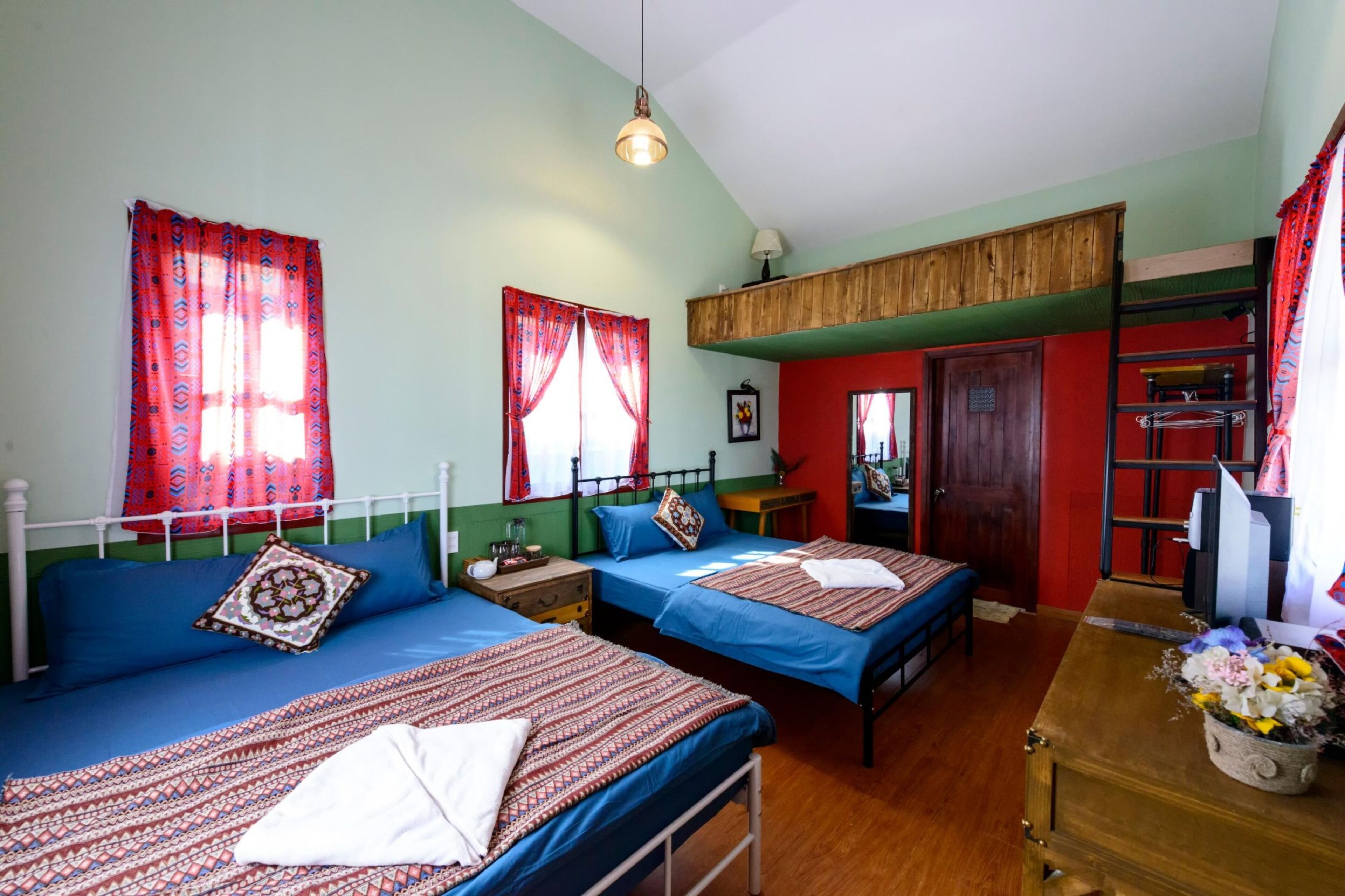 A room with two beds, a wooden dresser, a desk with a lamp, and a red door.