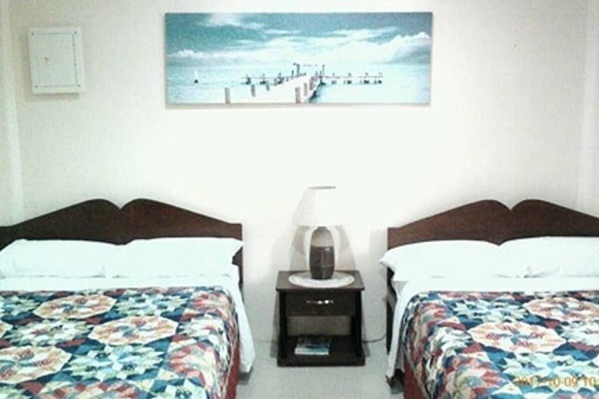 A hotel room with two beds, a nightstand with a lamp, and a painting of a pier on the wall.