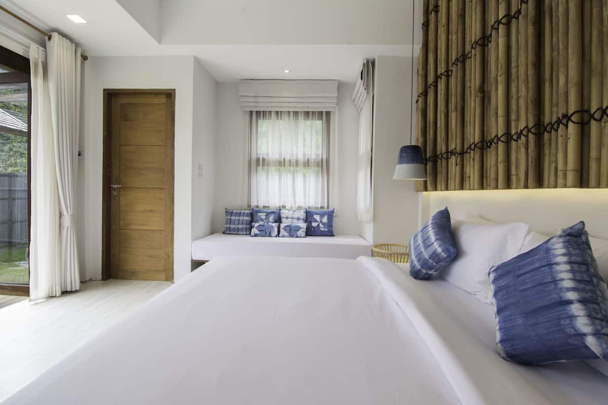 A bedroom with a large bed, a wooden door, a window with sheer curtains, and a bamboo wall.