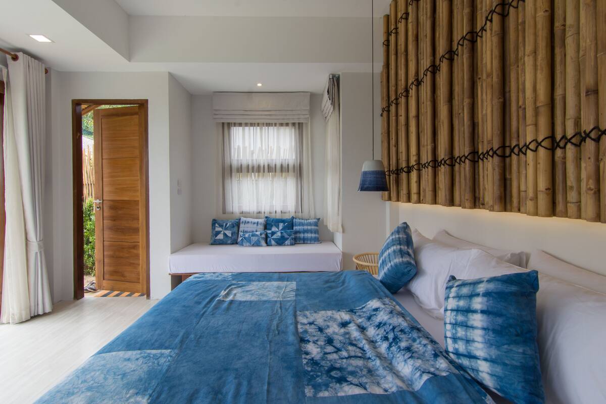 A bedroom with a large bed, a bedside table, a window with curtains, a wooden door, and a bamboo wall.