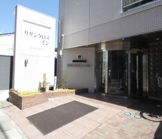 Entrada de hotel con puerta giratoria, un cartel con texto en japonés y un pequeño jardín con plantas.
