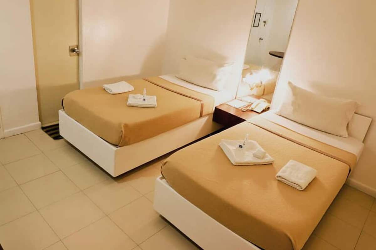 Une chambre d’hôtel avec deux lits simples, chacun doté d’un matelas blanc et de draps beiges. On y trouve une petite table de chevet avec une lampe et un livre. La chambre a un sol carrelé et une porte située sur le côté gauche.