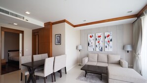 A modern living room with a dining area, a sofa, and a coffee table. There are three paintings on the wall, a lamp, and a door leading to a bedroom.