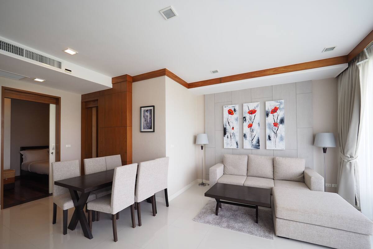Una sala moderna con comedor, un sofá y una mesa de centro. Hay tres cuadros en la pared, una lámpara y una puerta que da a un dormitorio.