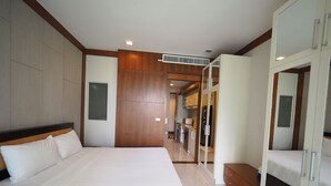 A modern hotel room with a large bed, a wooden wardrobe, and a kitchenette in the background.