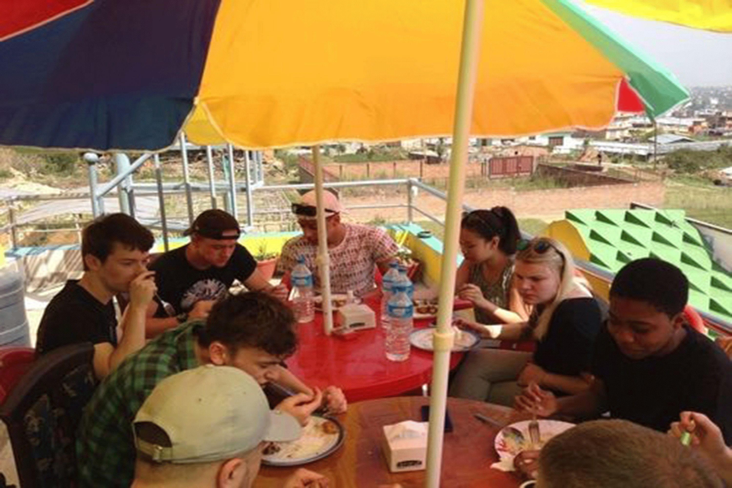 A group of people dining at an outdoor table with umbrellas, surrounded by a metal fence and greenery.