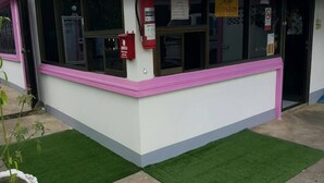 A building with a pink and white facade, a fire extinguisher mounted on the wall, and artificial green grass in front. - PKhotelresort (Prachin Buri)