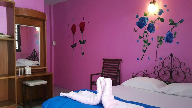 A bedroom with a pink wall, a bed with a blue and white bedspread, a wooden nightstand, a chair, and a wall-mounted light fixture. - PKhotelresort (Prachin Buri)