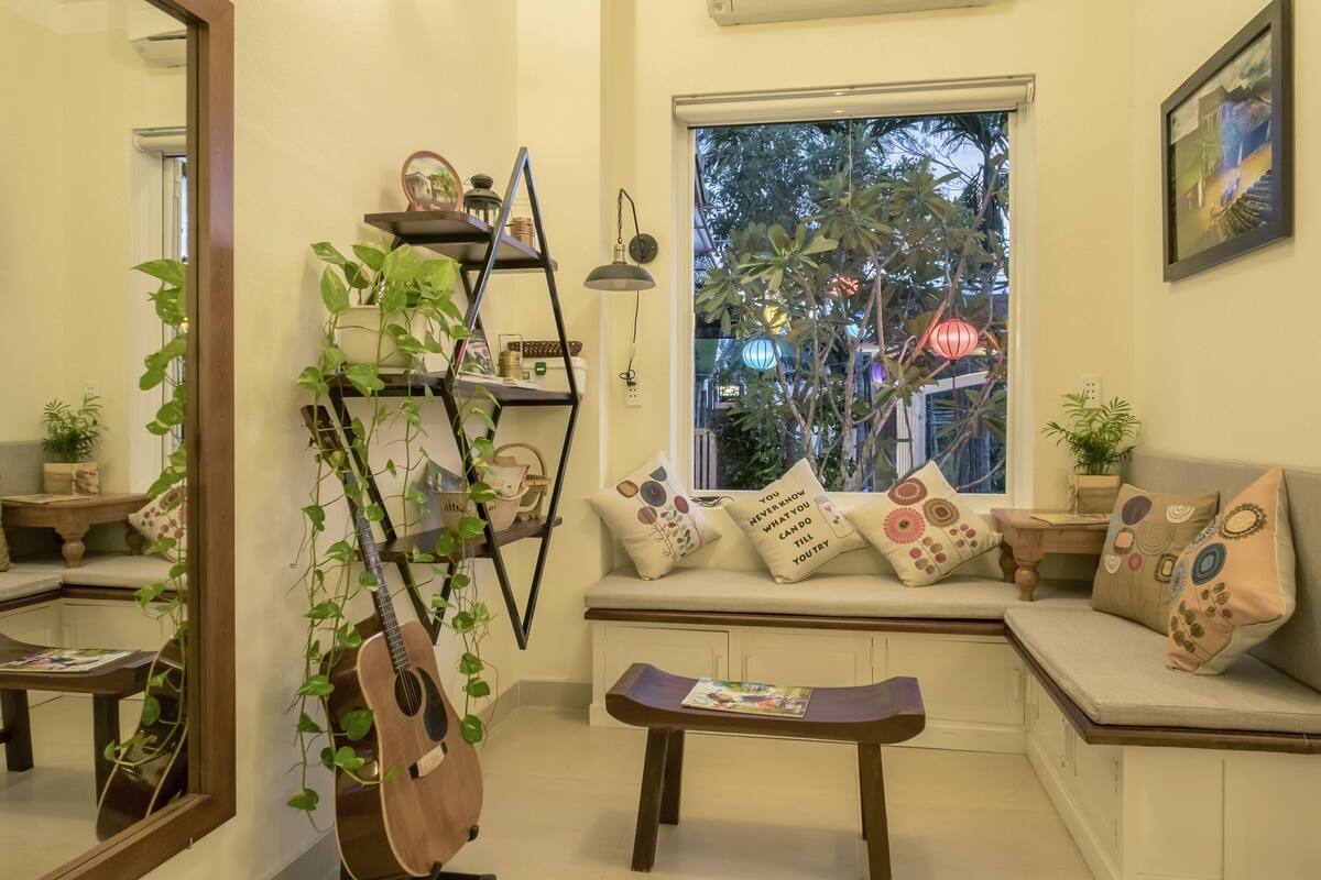 A cozy living room with a wooden guitar, a bookshelf, a window seat with cushions, and a small table.