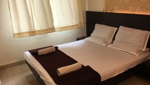 Una cama bien hecha con sábanas blancas y una colcha marrón oscuro, una mesita de noche con una lámpara y una ventana con cortinas traslúcidas.