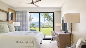 Une chambre d’hôtel équipée d’un lit, d’un bureau, d’une chaise, d’une télévision et d’une grande fenêtre donnant sur des palmiers et des collines.