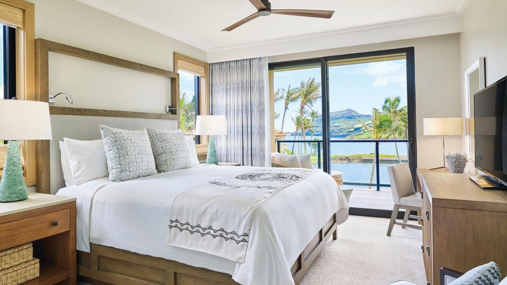 Habitación de hotel con una cama grande, un escritorio, televisor y un balcón con vista al agua y palmeras.