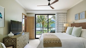 Une chambre d’hôtel avec un lit, un téléviseur posé sur une commode en bois, un ventilateur de plafond et une vue sur les palmiers et l’océan, visible à travers de grandes fenêtres.