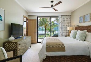 A hotel room with a bed, a television on a wooden dresser, a ceiling fan, and a view of palm trees and the ocean through large windows. - Timbers Kauai (Lihue)