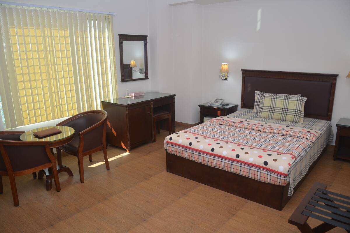 A hotel room with a wooden bed, a desk, a chair, a small table, a mirror, and a window with blinds.