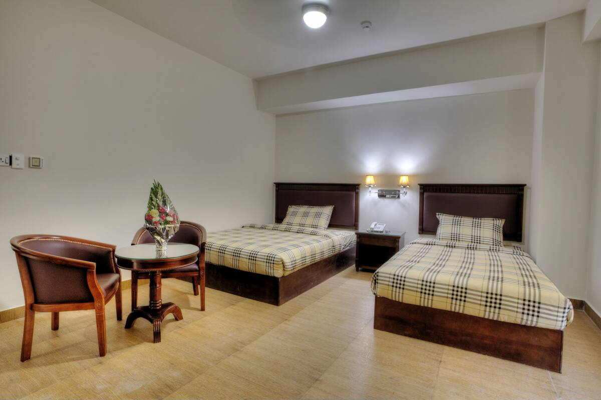 A hotel room with two beds, a chair, a small table with a vase, and a nightstand with a lamp.